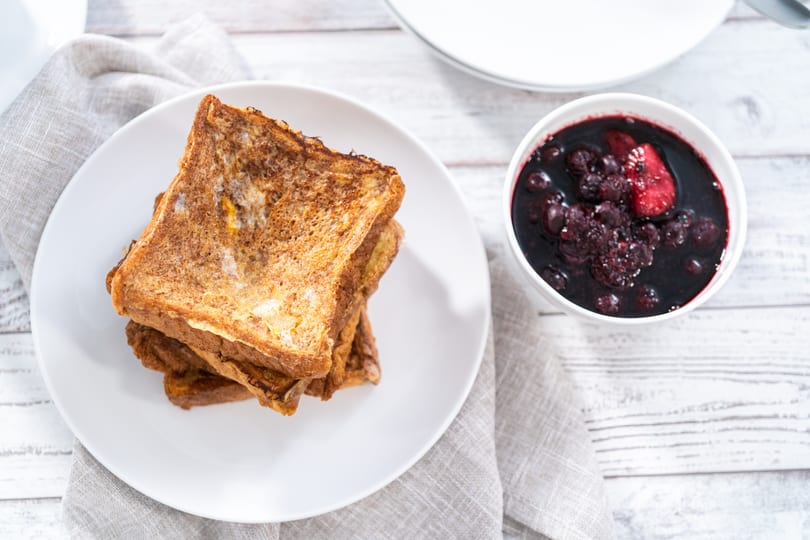 Golden brown French toast stacked on a white plate next to a bowl of dark berry compote.